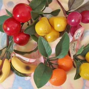 May include: A collection of artificial fruit, including red cherries, yellow pears, bananas, oranges, and lemons, with green leaves. Some pieces have tags that read "Made in British Hong Kong". The fruit is arranged on a floral patterned background.