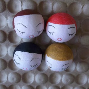 May include: Four handmade ceramic dolls with closed eyes and painted features. Each doll has a different hair color: brown, red, black, and blonde.
