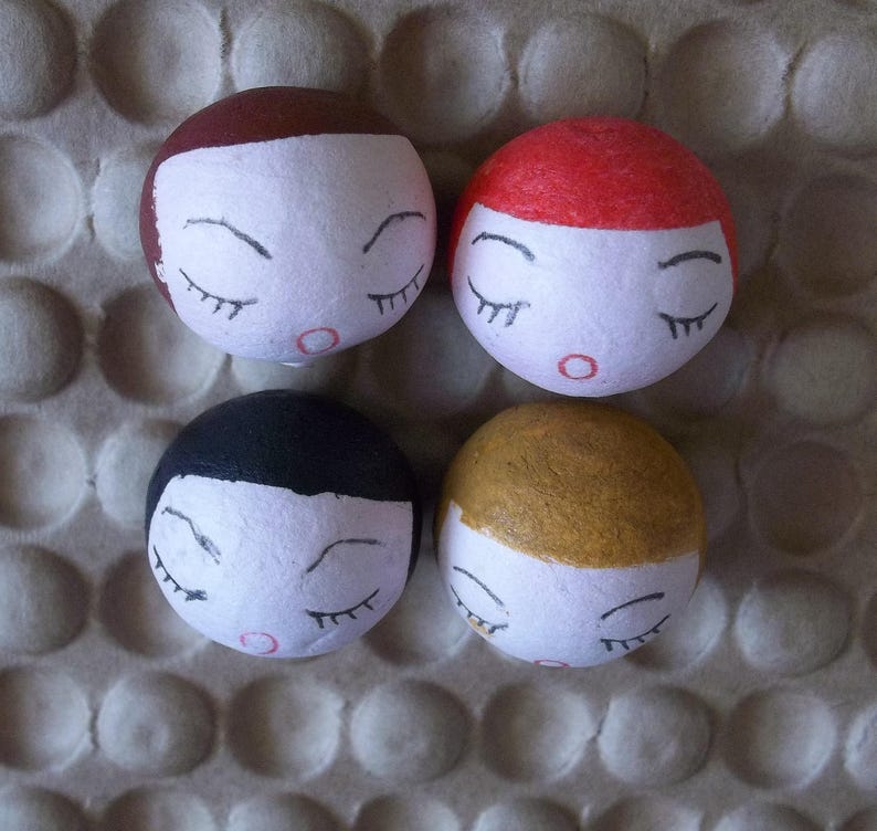 May include: Four handmade ceramic dolls with closed eyes and painted features. Each doll has a different hair color: brown, red, black, and blonde.