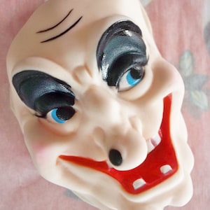 May include: A white plastic Halloween mask with a wide, toothy grin, black painted eyebrows, and black and blue painted eyes.