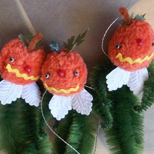 May include: Three hanging ornaments shaped like orange yarn pom-pom pumpkins with green pipe cleaner bodies, white fabric leaf collars, small black bead eyes, red bead noses, and yellow wavy ribbon mouths, each suspended by a thin silver string.