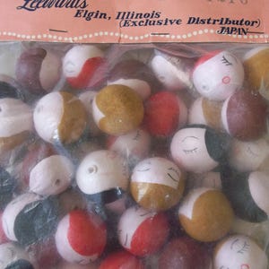 May include: A bag of assorted doll head beads. The beads are made of fabric and have painted faces. The beads are in various colors, including red, brown, white, and gold. The bag has a label that reads "Dexter brand Leewards Elgin, Illinois (Exclusive Distributor) JAPAN".