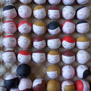 May include: A collection of small, round, painted ornaments with closed eyes and various facial expressions. The ornaments are arranged in a grid pattern on a white background.