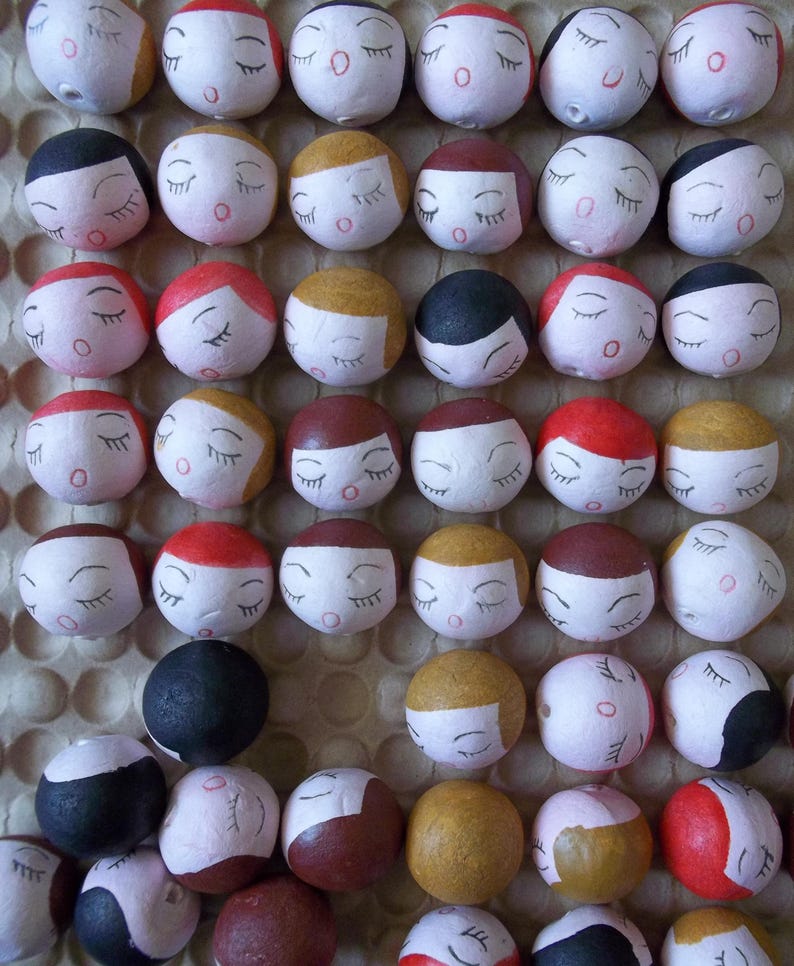 May include: A collection of small, round, painted ornaments with closed eyes and various facial expressions. The ornaments are arranged in a grid pattern on a white background.