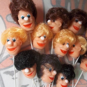 May include: A collection of vintage clown heads on sticks. The clown heads have various hair colors, including brown, blonde, and black. Each head features a white nose, blue eyes, and a red smile. The heads are made of plastic.