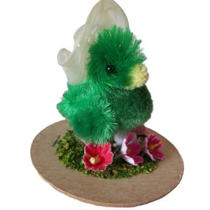 May include: A handmade, plush green duck figurine with a yellow beak and black eyes. The duck is perched on a round, brown base with green moss and red and pink flowers. A sheer, cream-colored veil is attached to the duck's head.
