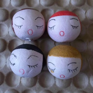 May include: Four round, white, styrofoam ornaments with painted faces. Each ornament has black eyes, eyelashes, and a red mouth. The ornaments have different colored accents on top: brown, red, gold, and black.