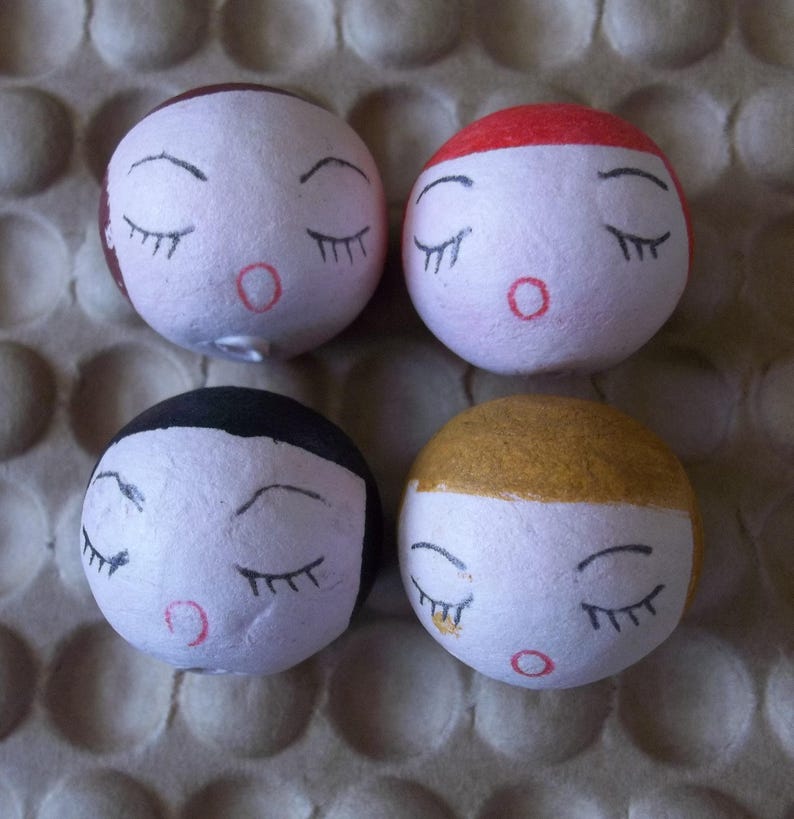 May include: Four round, white, styrofoam ornaments with painted faces. Each ornament has black eyes, eyelashes, and a red mouth. The ornaments have different colored accents on top: brown, red, gold, and black.