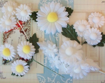 May include: Assortment of white fabric daisy flowers with yellow centers. The flowers are arranged on a white background with blue and white patterned paper.