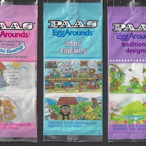 May include: Three packages of PAAS EggArounds Easter egg decorating kits. The pink package features a design with two girls under an umbrella and the text "Special Blessings". The blue package features a design with a bunny rabbit and the text "PAAS FRIENDS". The purple package features a design with a rainbow and the text "traditional designs".