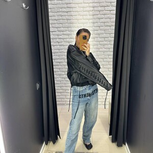 Op de afbeelding: Een persoon draagt een zwarte jas met vetersluiting op de mouwen en een lichtblauwe denim jeans. Op de jeans staat het woord "VETEMENTS" gedrukt. De persoon bevindt zich in een paskamer met zwarte gordijnen en een witte bakstenen muur.
