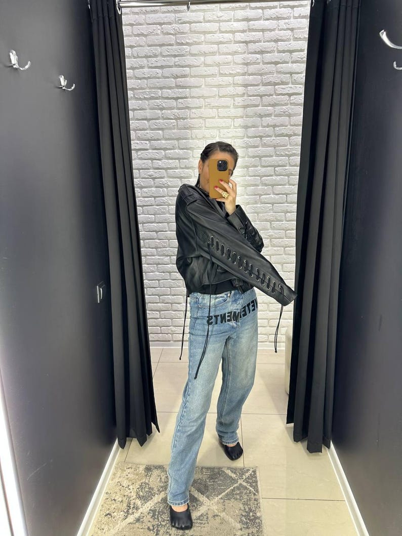 Op de afbeelding: Een persoon draagt een zwarte jas met vetersluiting op de mouwen en een lichtblauwe denim jeans. Op de jeans staat het woord "VETEMENTS" gedrukt. De persoon bevindt zich in een paskamer met zwarte gordijnen en een witte bakstenen muur.