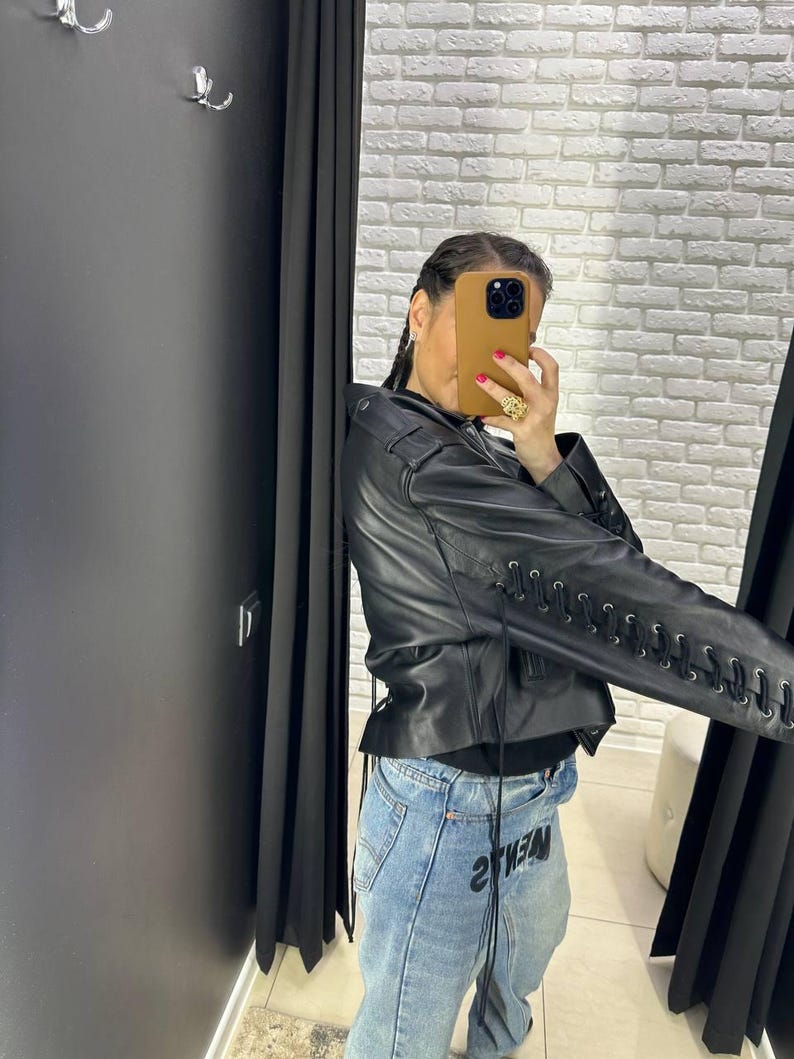 Op de afbeelding: Zwarte leren jas met vetersluiting op de mouwen. De jas heeft een klassieke bikerstijl met een reverskraag en een getailleerd silhouet. Het model draagt een blauwe jeans met het woord "2THEM" op de voorkant.