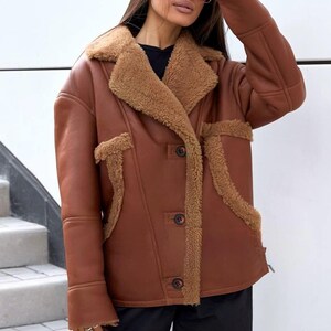Puede incluir: Chaqueta marr&oacute;n de piel sint&eacute;tica con cuello y ribete de borrego. La chaqueta tiene cierre de botones y dos bolsillos delanteros. La modelo lleva gafas de sol y pantalones negros. Prenda de abrigo c&aacute;lida y elegante.