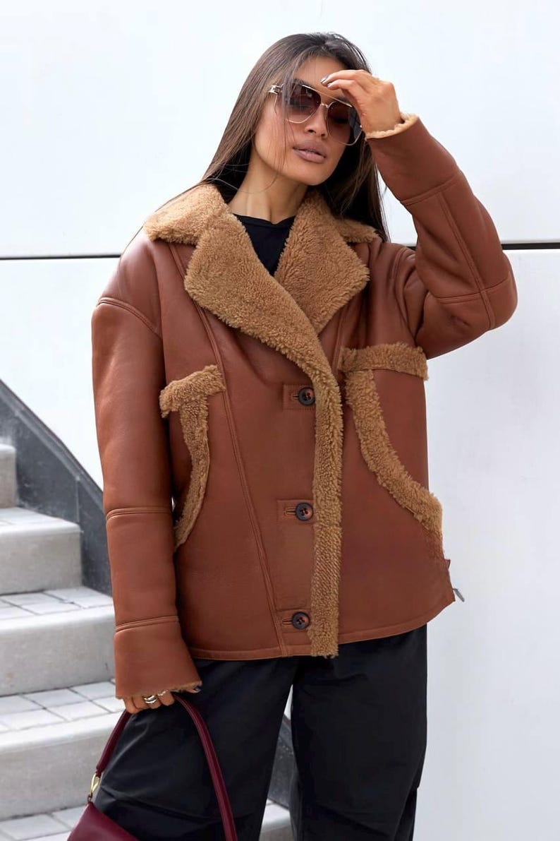 Puede incluir: Chaqueta marr&oacute;n de piel sint&eacute;tica con cuello y ribete de borrego. La chaqueta tiene cierre de botones y dos bolsillos delanteros. La modelo lleva gafas de sol y pantalones negros. Prenda de abrigo c&aacute;lida y elegante.