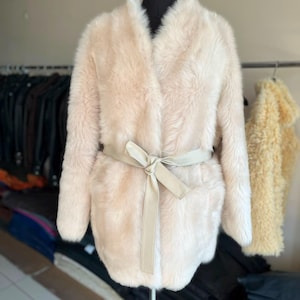 May include: A cream-colored faux fur coat featuring long sleeves, an open front design, and a matching cream-colored smooth fabric tie belt that cinches at the waist. The coat has a soft, plush texture and reaches mid-thigh length on the mannequin.