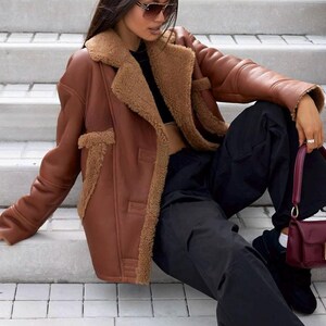 Puede incluir: Chaqueta marr&oacute;n de cuero sint&eacute;tico con forro y ribete de borreguillo. La chaqueta tiene una parte delantera abotonada y se combina con pantalones negros y un bolso burdeos. La modelo lleva gafas de sol y est&aacute; sentada en escalones de hormig&oacute;n.