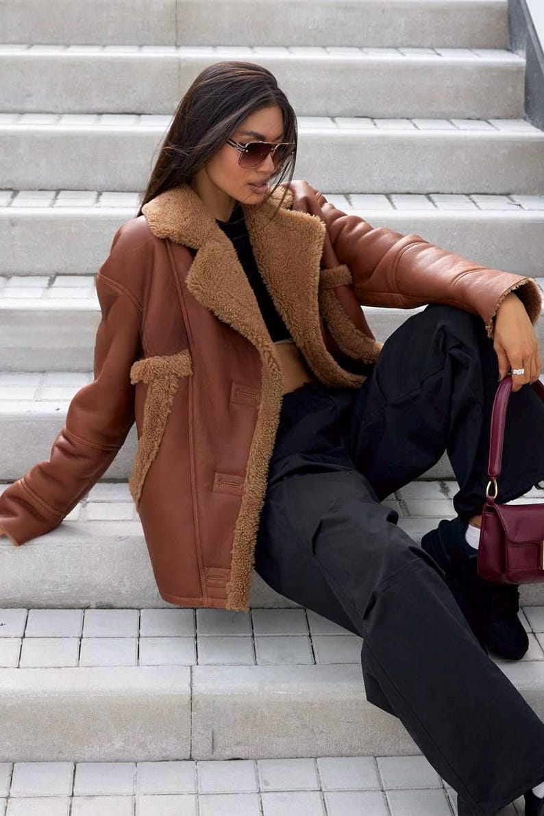 Puede incluir: Chaqueta marr&oacute;n de cuero sint&eacute;tico con forro y ribete de borreguillo. La chaqueta tiene una parte delantera abotonada y se combina con pantalones negros y un bolso burdeos. La modelo lleva gafas de sol y est&aacute; sentada en escalones de hormig&oacute;n.