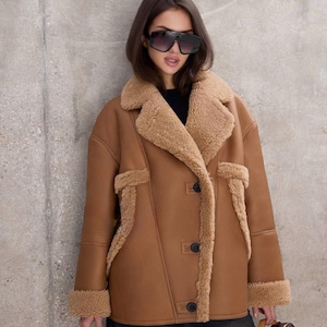 Puede incluir: Chaqueta de borrego marr&oacute;n con forro y ribete de piel sint&eacute;tica beige. La chaqueta tiene cierre de botones y dos bolsillos. La persona lleva gafas de sol negras y vaqueros oscuros. Una opci&oacute;n de abrigo elegante.