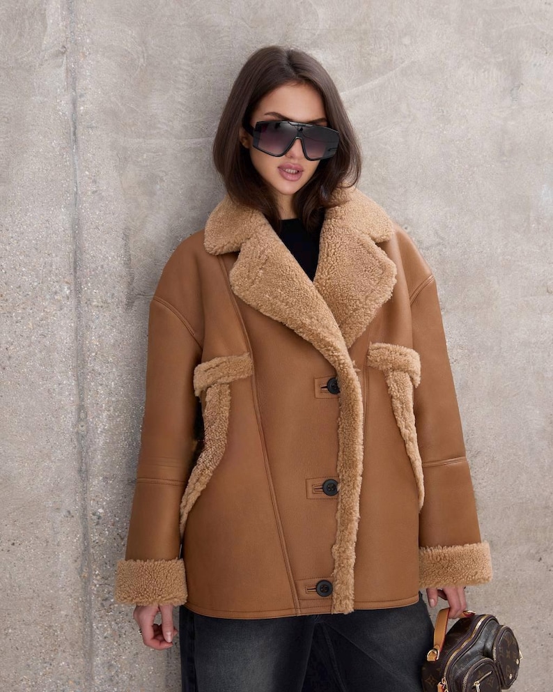 Puede incluir: Chaqueta de borrego marr&oacute;n con forro y ribete de piel sint&eacute;tica beige. La chaqueta tiene cierre de botones y dos bolsillos. La persona lleva gafas de sol negras y vaqueros oscuros. Una opci&oacute;n de abrigo elegante.