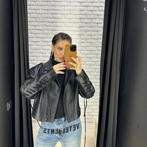 Op de afbeelding: Zwarte leren jas met vetersluiting op de mouwen. De jas wordt gecombineerd met een lichtblauwe jeans met het woord "VETEMENTS" in omgekeerde volgorde. De persoon houdt een telefoon in een bruine hoes vast.