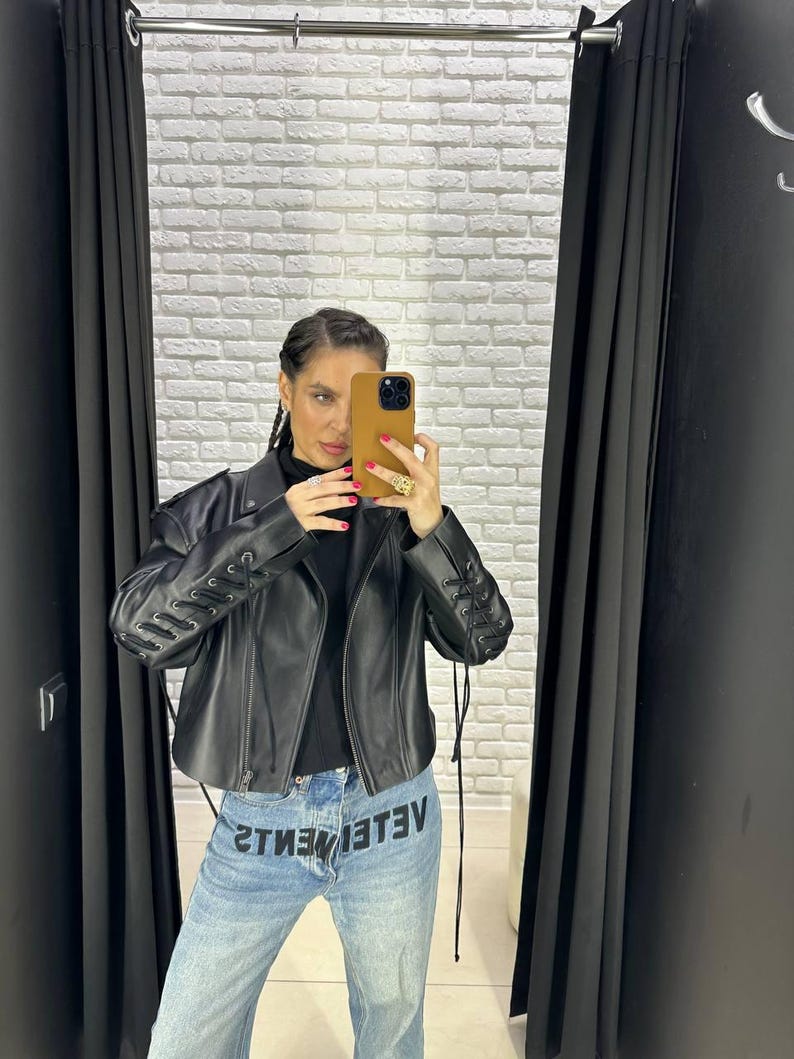 Op de afbeelding: Zwarte leren jas met vetersluiting op de mouwen. De jas wordt gecombineerd met een lichtblauwe jeans met het woord "VETEMENTS" in omgekeerde volgorde. De persoon houdt een telefoon in een bruine hoes vast.