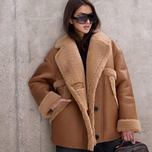 Puede incluir: Un abrigo de borrego marr&oacute;n con interior beige y solapas grandes. El abrigo tiene cierre de botones y se lleva con pantalones negros y gafas de sol. Un bolso marr&oacute;n con un logotipo se sostiene en la mano.