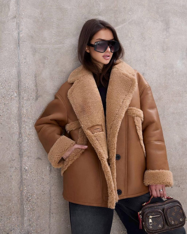 Puede incluir: Un abrigo de borrego marr&oacute;n con interior beige y solapas grandes. El abrigo tiene cierre de botones y se lleva con pantalones negros y gafas de sol. Un bolso marr&oacute;n con un logotipo se sostiene en la mano.