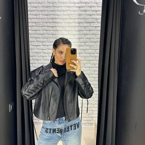 Op de afbeelding: Zwarte leren jas met vetersluiting op de mouwen. De jas wordt gecombineerd met een lichtblauwe denim jeans met de zwarte tekst "VETEMENTS". De persoon draagt een zwarte coltrui.