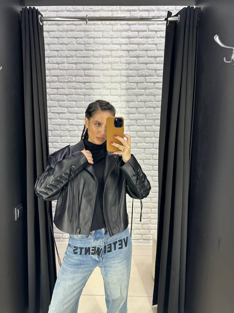 Op de afbeelding: Zwarte leren jas met vetersluiting op de mouwen. De jas wordt gecombineerd met een lichtblauwe denim jeans met de zwarte tekst "VETEMENTS". De persoon draagt een zwarte coltrui.