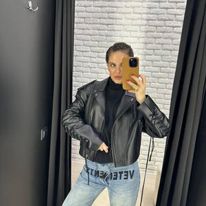 Op de afbeelding: Een zwarte leren jas met vetersluiting op de mouwen. De jas wordt gecombineerd met een lichtblauwe denim jeans met het woord "VETEMENTS" in het zwart gedrukt. De persoon draagt een zwarte coltrui.