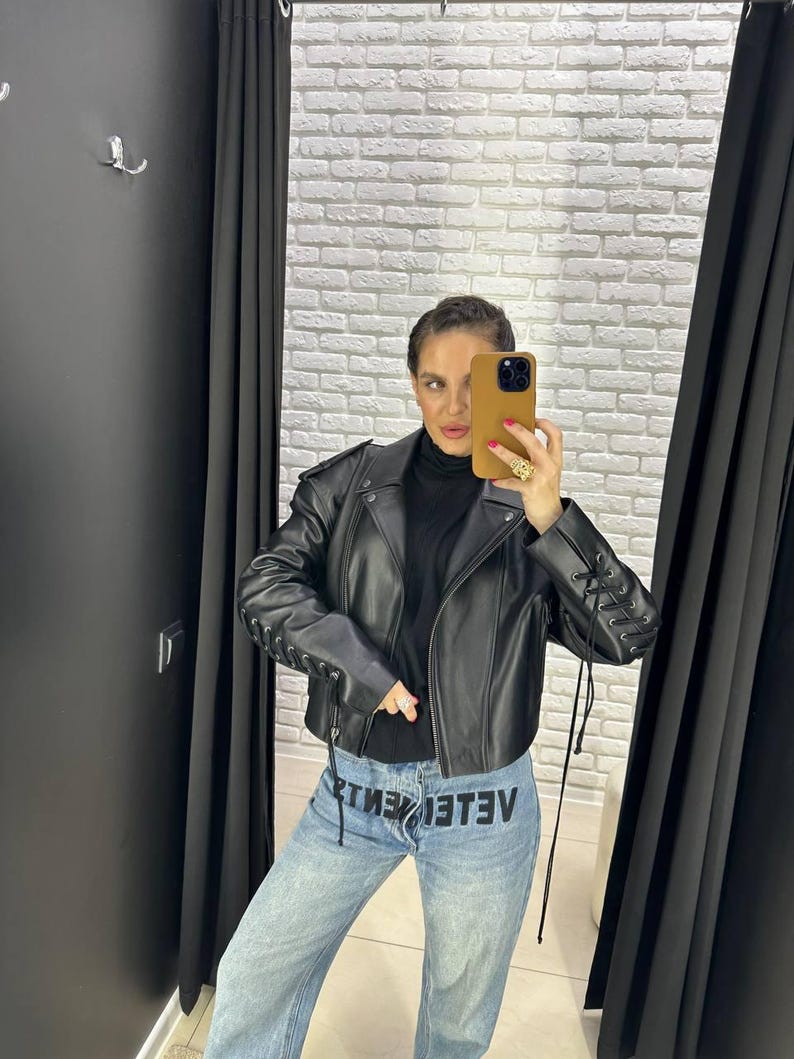 Op de afbeelding: Een zwarte leren jas met vetersluiting op de mouwen. De jas wordt gecombineerd met een lichtblauwe denim jeans met het woord "VETEMENTS" in het zwart gedrukt. De persoon draagt een zwarte coltrui.
