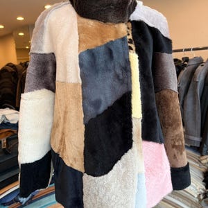 May include: A patchwork coat in various colors, including black, brown, beige, and pink, is displayed on a mannequin. The coat features a high, dark brown collar and a mix of textures. The design is a unique blend of geometric shapes and colors.