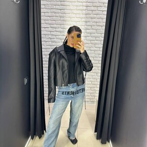 Op de afbeelding: Een zwarte leren jasje met een cropped design en een zwarte coltrui. Het model draagt lichtblauwe jeans met het woord "VETEMENTS" op de voorkant. De achtergrond is een witte bakstenen muur en zwarte gordijnen.