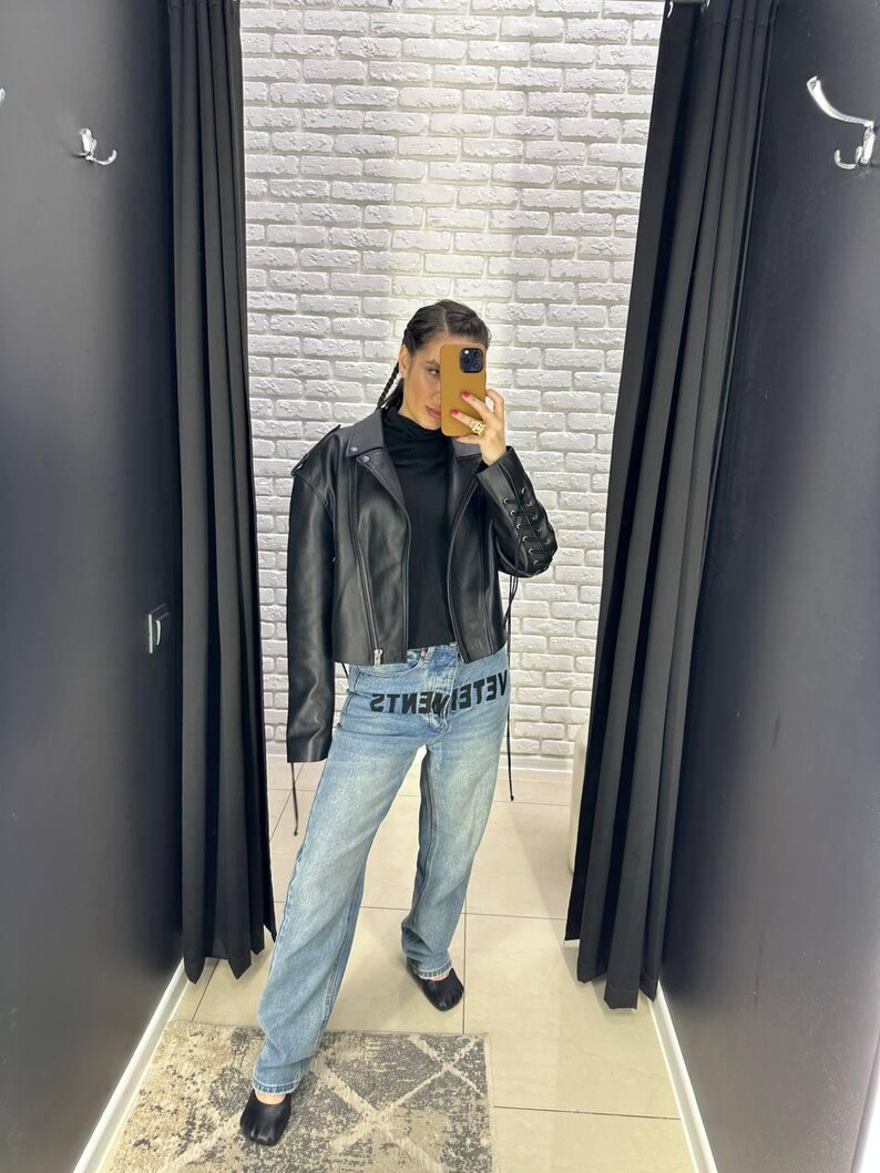 Op de afbeelding: Een zwarte leren jasje met een cropped design en een zwarte coltrui. Het model draagt lichtblauwe jeans met het woord "VETEMENTS" op de voorkant. De achtergrond is een witte bakstenen muur en zwarte gordijnen.