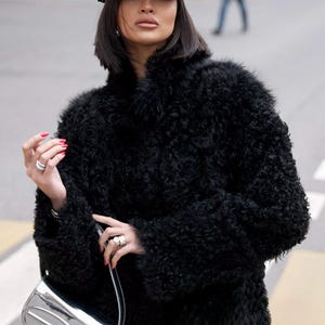 May include: A black baseball cap with the white text Miu Miu, a black textured faux fur coat with long sleeves, and a metallic silver shoulder bag with a prominent embossed logo on the front. The accessories include silver rings on the fingers.