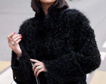 May include: A black baseball cap with the white text Miu Miu, a black textured faux fur coat with long sleeves, and a metallic silver shoulder bag with a prominent embossed logo on the front. The accessories include silver rings on the fingers.