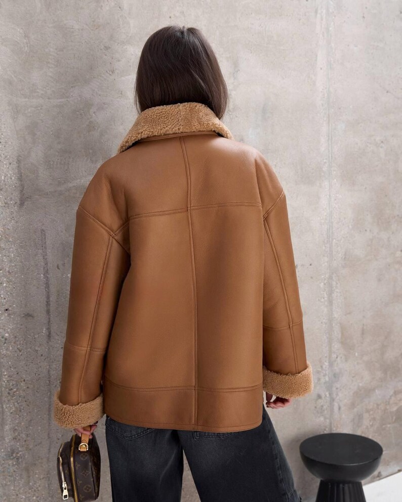 Puede incluir: Chaqueta de borreguillo marr&oacute;n con cuello y pu&ntilde;os de borreguillo beige. La chaqueta tiene un dise&ntilde;o cl&aacute;sico con costuras visibles y un corte relajado. El modelo lleva vaqueros oscuros.