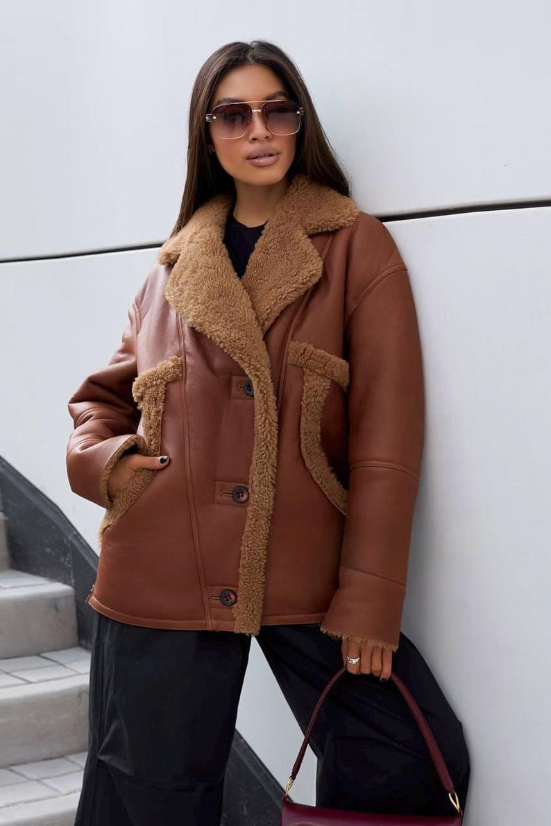 Puede incluir: Chaqueta de cuero marr&oacute;n con cuello y ribetes de borrego. La chaqueta tiene cierre de botones y dos bolsillos delanteros. La modelo lleva pantalones negros y gafas de sol.