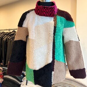 May include: A patchwork jacket in a variety of colors, including black, white, tan, green, and brown. The jacket has a high, pink and black patterned collar and is buttoned up the front. The jacket is displayed on a mannequin.