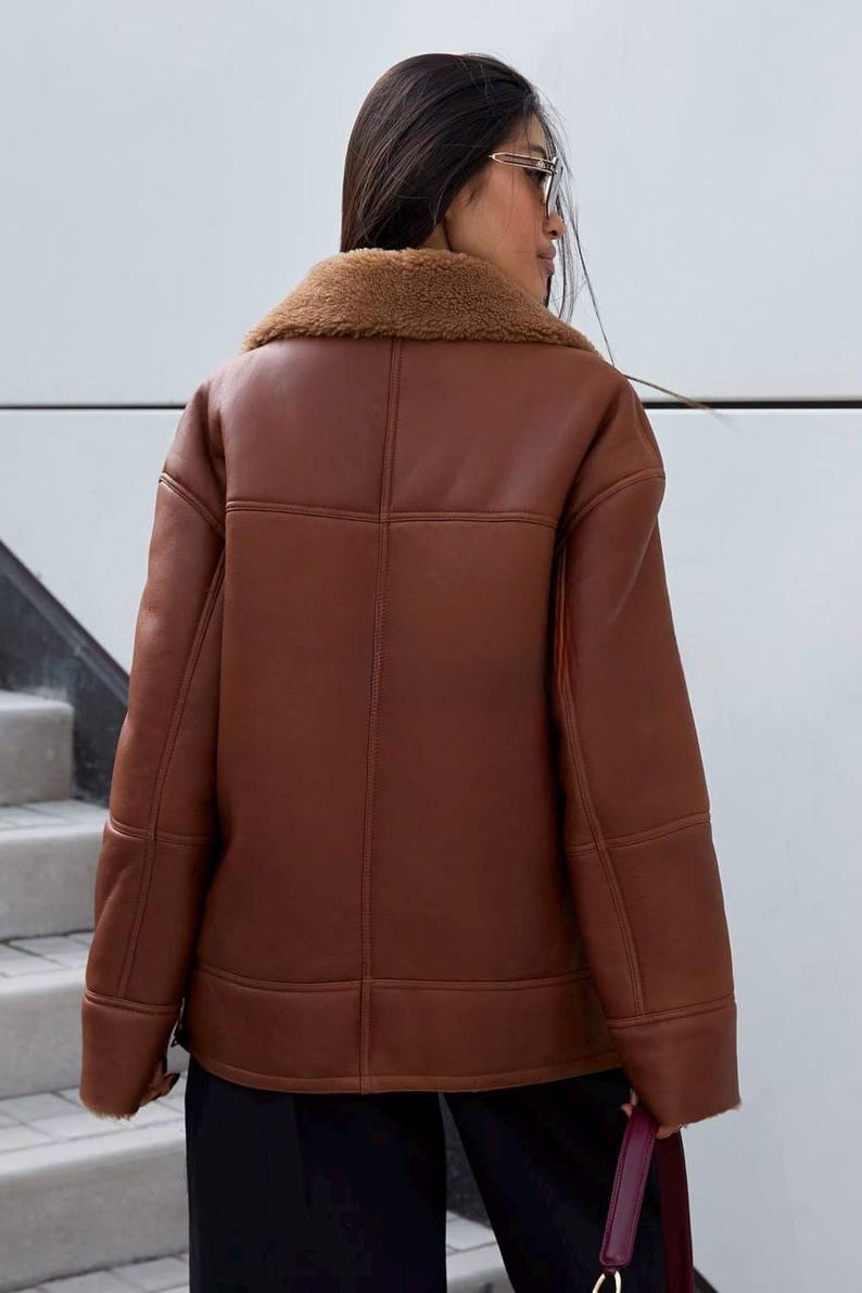 Puede incluir: Chaqueta de cuero marr&oacute;n con cuello de borrego. La chaqueta tiene un corte holgado y presenta detalles de costura visibles. El modelo lleva la chaqueta con pantalones negros. Una opci&oacute;n de abrigo c&aacute;lida y elegante.