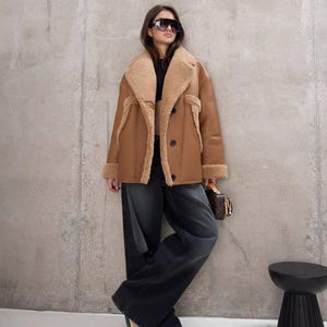 Puede incluir: Un abrigo de borrego marr&oacute;n con interior y ribetes color beige. El abrigo est&aacute; abotonado y se lleva con vaqueros de mezclilla oscura y zapatillas deportivas. La persona lleva gafas de sol y sostiene un peque&ntilde;o bolso marr&oacute;n. Hay un taburete negro al lado.