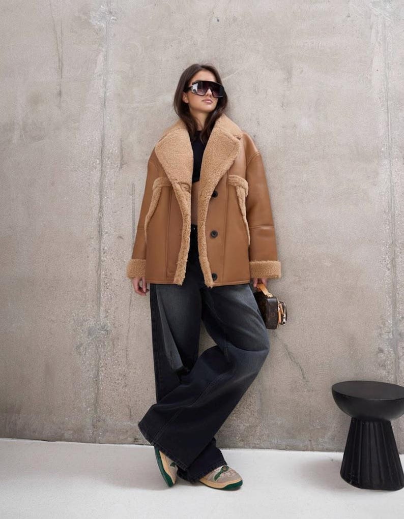 Puede incluir: Un abrigo de borrego marr&oacute;n con interior y ribetes color beige. El abrigo est&aacute; abotonado y se lleva con vaqueros de mezclilla oscura y zapatillas deportivas. La persona lleva gafas de sol y sostiene un peque&ntilde;o bolso marr&oacute;n. Hay un taburete negro al lado.