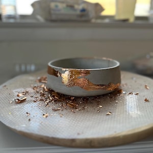 Puede incluir: Un pequeño cuenco de hormigón gris con una banda de pan de oro en el centro. Copos de pan de oro están esparcidos alrededor del cuenco sobre una superficie texturizada blanca y dorada. El cuenco es probablemente hecho a mano.