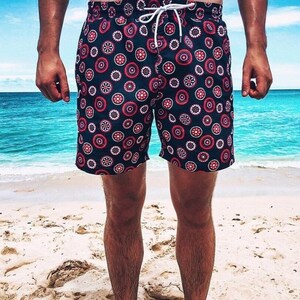 May include: Navy blue swim trunks with a repeating pattern of red, white, and navy blue circular designs. The shorts have a drawstring closure and are worn on a sandy beach with a turquoise ocean in the background.