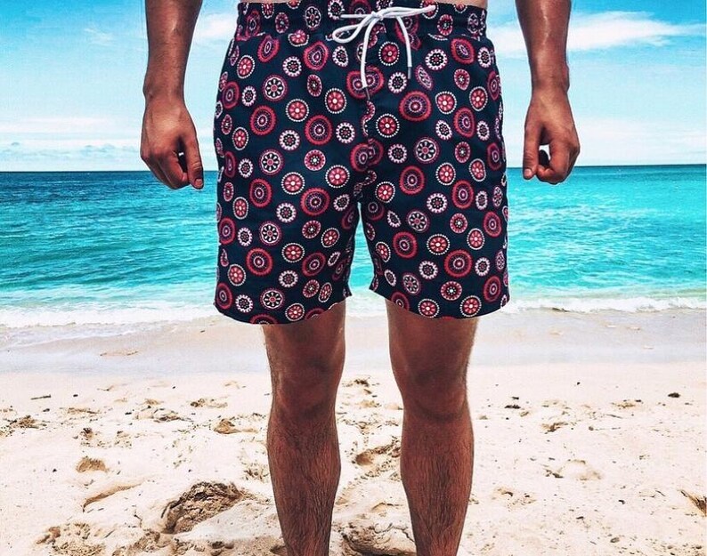 May include: Navy blue swim trunks with a repeating pattern of red, white, and navy blue circular designs. The shorts have a drawstring closure and are worn on a sandy beach with a turquoise ocean in the background.