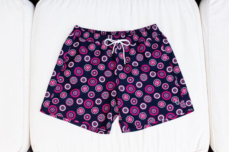 May include: Navy blue swim shorts with a repeating pattern of pink and white circular designs. The shorts have a white drawstring and are made of a lightweight fabric. The shorts are designed for swimming and other water activities.