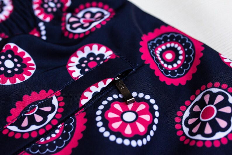 May include: Close-up of navy blue fabric with a repeating paisley pattern in pink, white, and black. A small pocket with a metal zipper is visible. The fabric has a slight sheen.