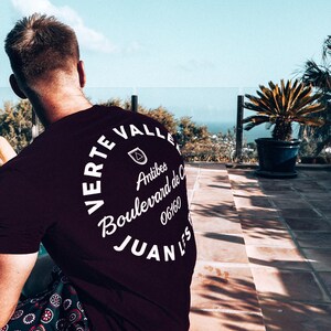 May include: A dark burgundy t-shirt with white text in a circular design. The text reads "Verte Valle Antibes Boulevard de C 06160 Juan Les". The person is wearing patterned shorts. A potted plant and a view of the ocean are in the background.