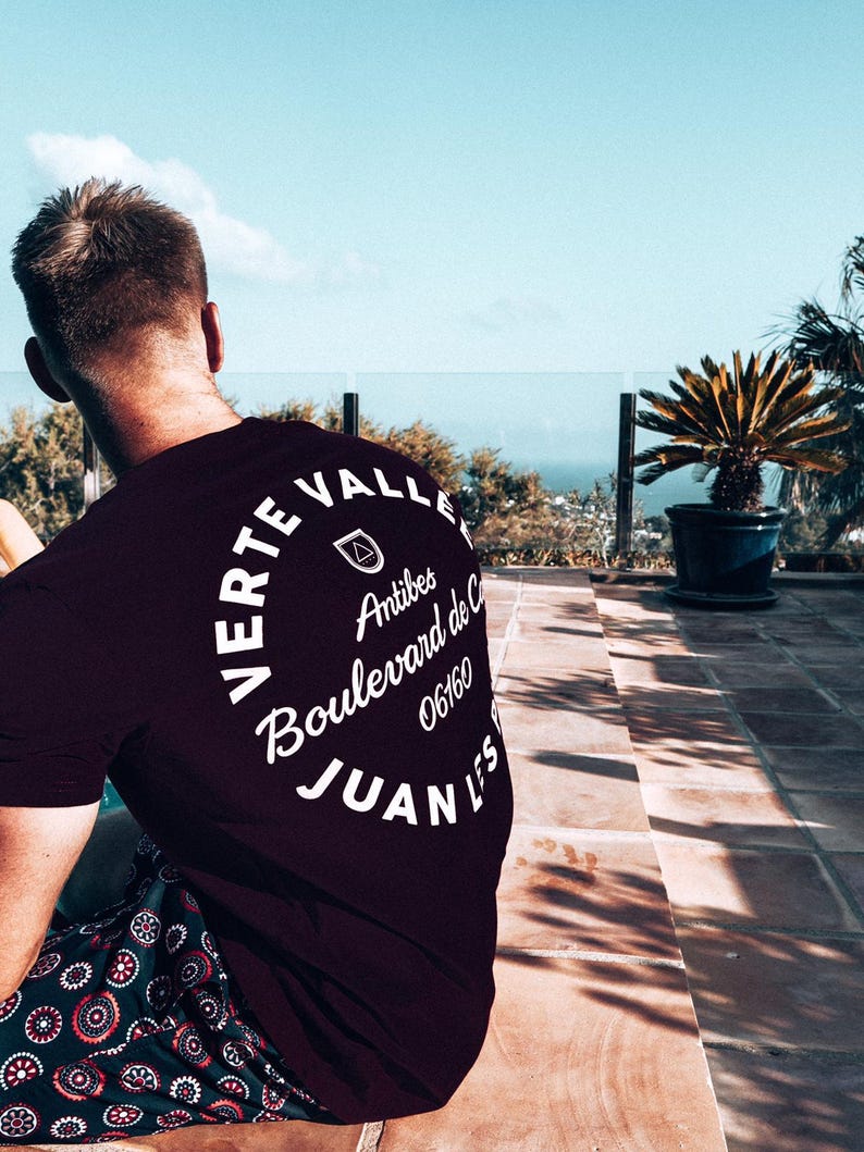 May include: A dark burgundy t-shirt with white text in a circular design. The text reads "Verte Valle Antibes Boulevard de C 06160 Juan Les". The person is wearing patterned shorts. A potted plant and a view of the ocean are in the background.